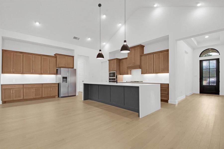 Virtually-Staged Kitchen with wood cabinetry, quartz countertops, 16ft vaulted ceiling, a kitchen breakfast bar, pendant lighting, a center island with sink, stainless steel appliances, and light hardwood floors
