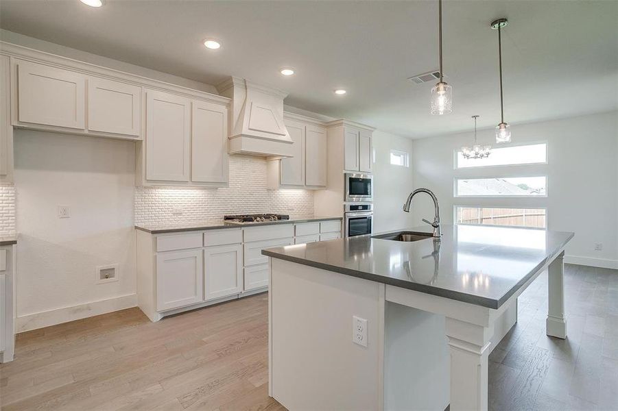 Kitchen featuring custom range hood, light hardwood / wood-style floors, backsplash, a center island with sink, and white cabinetry