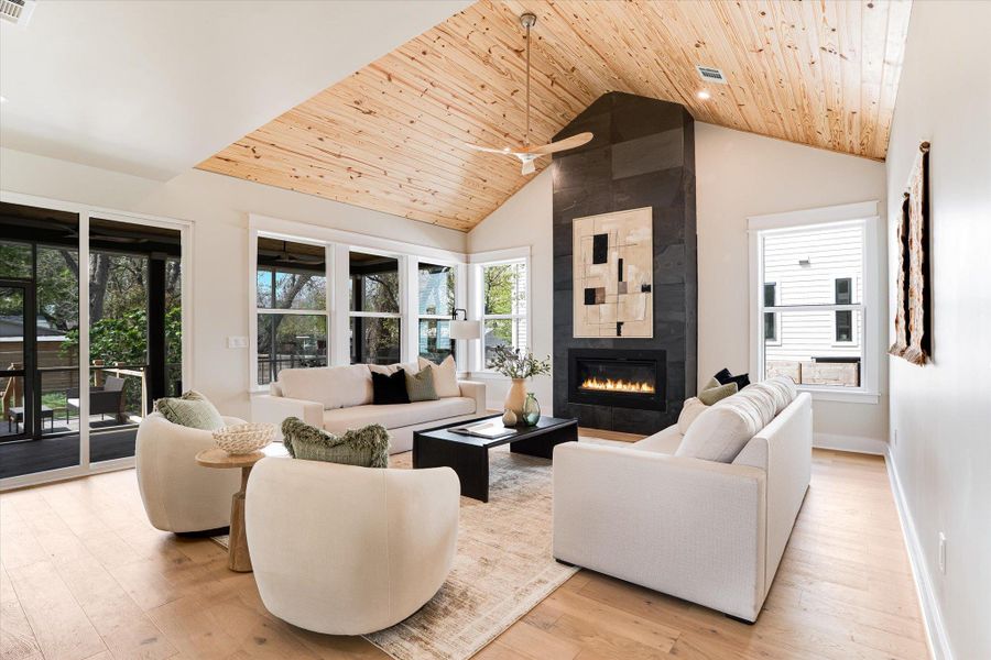 This living space features a vaulted wood plank ceiling, light wood flooring, an impressive fireplace with a dark stone surround and contemporary art piece, and large windows that provide natural light This living space features a vaulted wood plank ceiling, light wood flooring, an impressive fireplace with a dark stone surround and contemporary art piece, and large windows that provide natural light