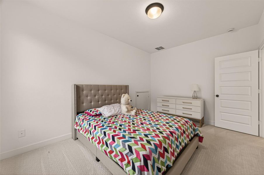 This bedroom features a cozy bed with a colorful chevron-patterned blanket, a plush headboard, and a small stuffed animal. The room is complemented by a sleek dresser, a nightstand with a lamp, and a simple yet modern ceiling light. The neutral walls and carpet provide a clean, inviting atmosphere.
