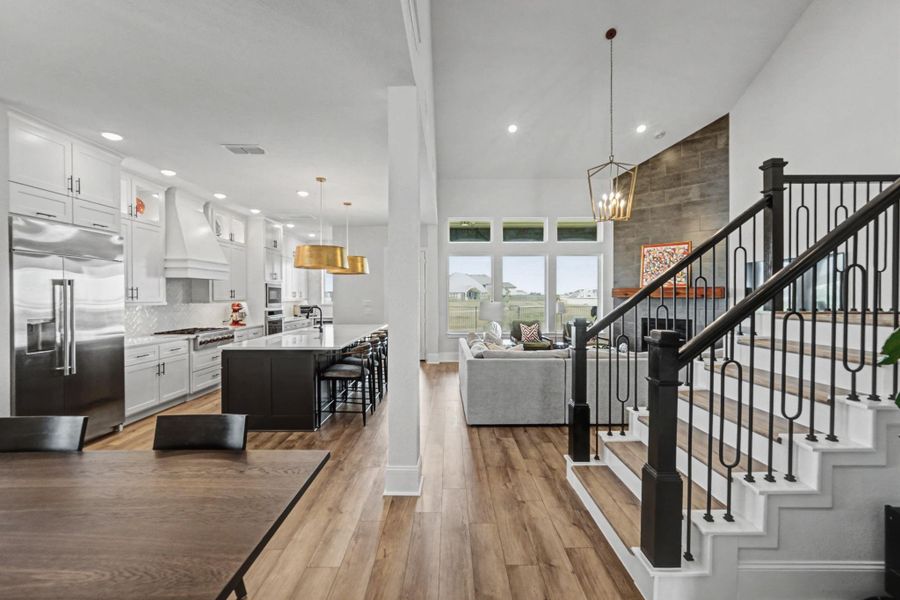 This wide view captures the seamless connection between the kitchen, the open living area with its dramatic floor-to-ceiling stone fireplace, and a stylish staircase. Vinyl plank flooring runs throughout, uniting the distinct spaces.
