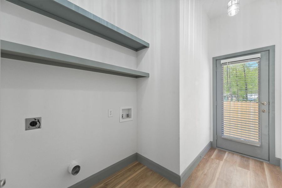 Bright laundry room with hookups for washer and dryer, featuring built-in shelving. A door with glass panels and blinds leads outside, providing natural light and easy access.