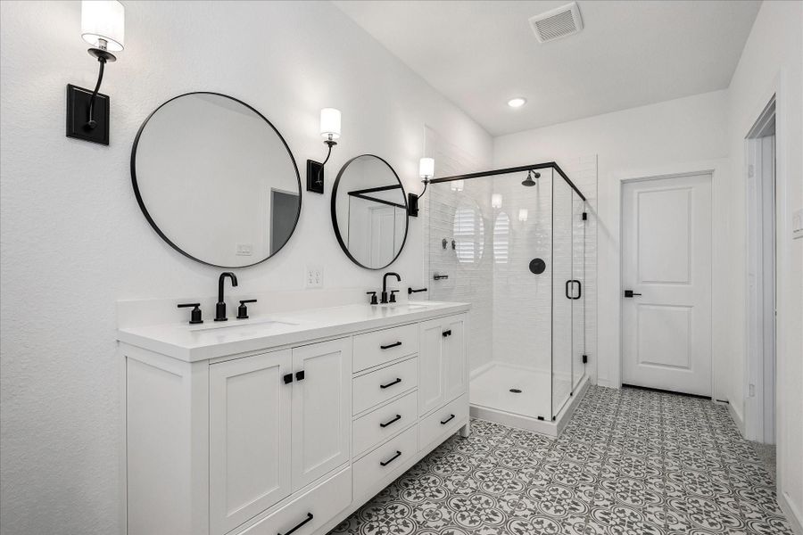 Designer primary bath featuring dual vanities, statement round mirrors, patterned tile flooring, and a frameless glass walk-in shower.