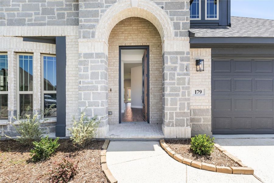 Stone and brick home entrance with arched doorway, open front door, and attached garage beside landscaped walkway Stone and brick home entrance with arched doorway, open front door, and attached garage beside landscaped walkway