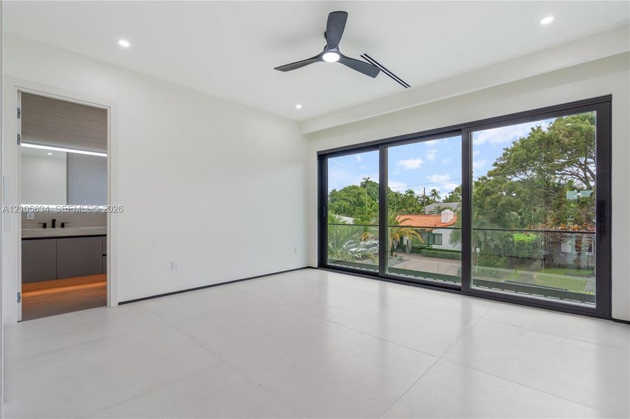 Large-format porcelain tile flooring, expansive floor-to-ceiling sliding glass doors with black frames, recessed LED lighting, three-blade ceiling fan, and balcony with glass railing