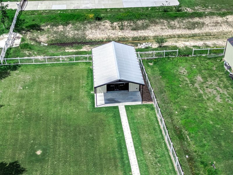 This aerial view highlights the fenced barn-style structure positioned on the expansive grassy portion of the property, offering additional versatility for storage, hobbies, equipment, recreational use, or future customization.