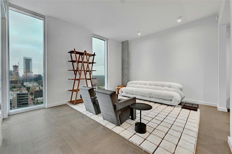 A minimalist design, featuring light-colored wood flooring arranged in a herringbone pattern. The room is brightly lit by natural light streaming in through large, floor-to-ceiling glass windows and door that lead to a balcony. *Linden Residence No. 2406 Furnished Model Shown Here (Furnishings provided by Scott + Cooner / Cassina Available for Purchase)