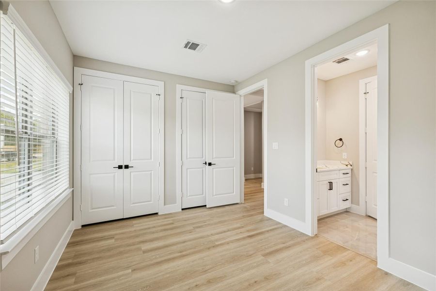 This bright room features light wood flooring, a large window with blinds, and a double-door closet. It connects to a bathroom with modern fixtures, offering a convenient and functional layout.