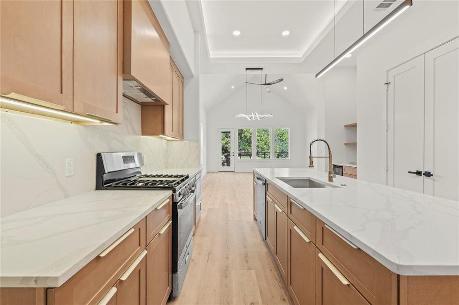 Kitchen with light wood-style flooring, decorative light fixtures, appliances with stainless steel finishes, light stone counters, and a center island with sink Kitchen with light wood-style flooring, decorative light fixtures, appliances with stainless steel finishes, light stone counters, and a center island with sink