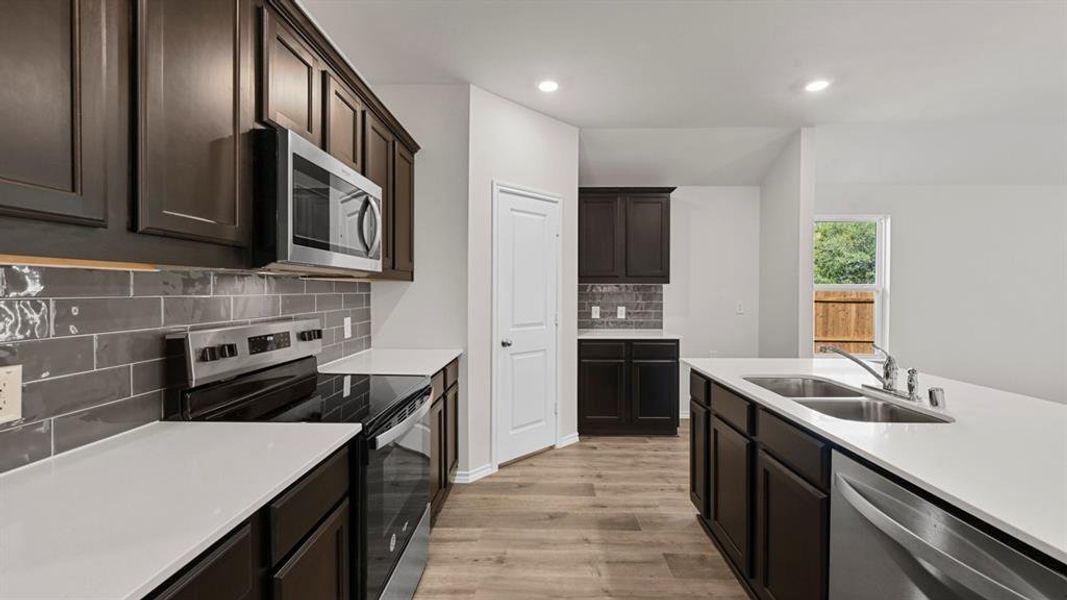 Modern kitchen featuring dark wood-finish cabinetry, stainless steel appliances, a subway tile backsplash, light countertops, and wood-finish flooring Modern kitchen featuring dark wood-finish cabinetry, stainless steel appliances, a subway tile backsplash, light countertops, and wood-finish flooring