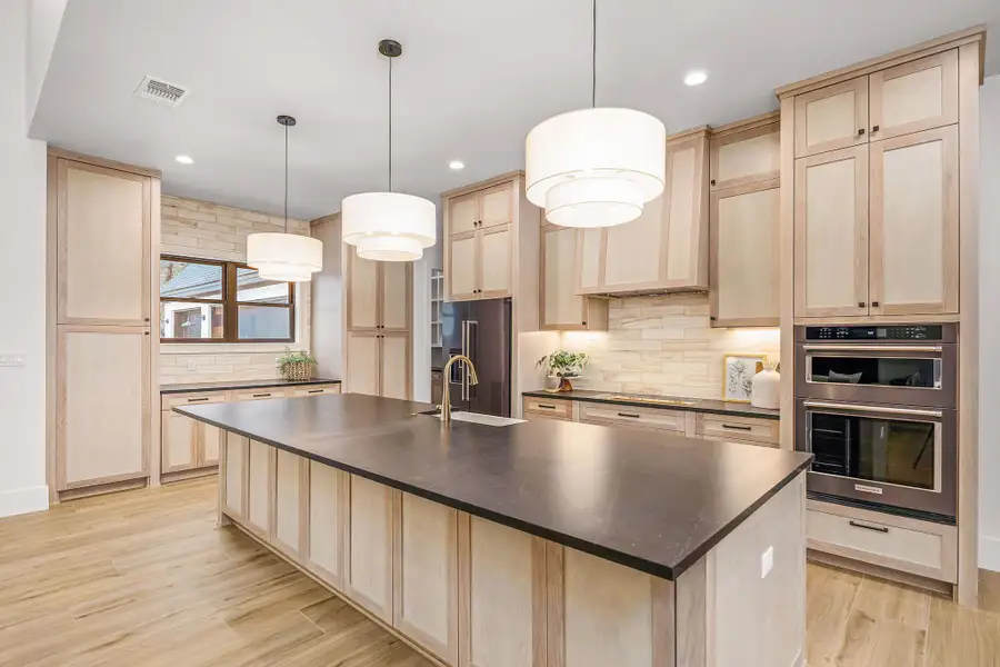 Kitchen featuring dark countertops, decorative light fixtures, a center island with sink, and light brown cabinets