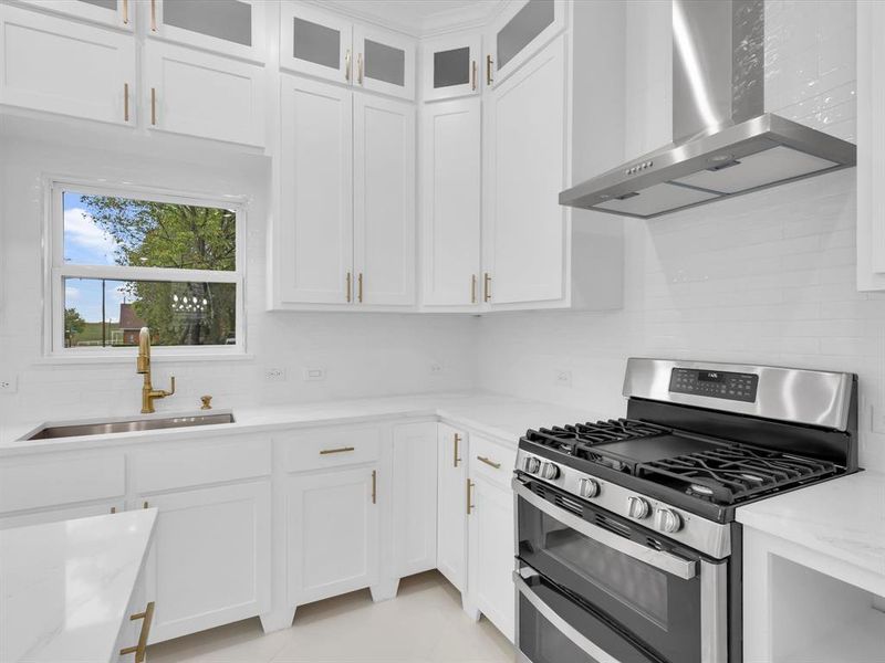 Kitchen featuring light stone counters, white cabinetry, stainless steel gas range oven, sink, and wall chimney exhaust hood