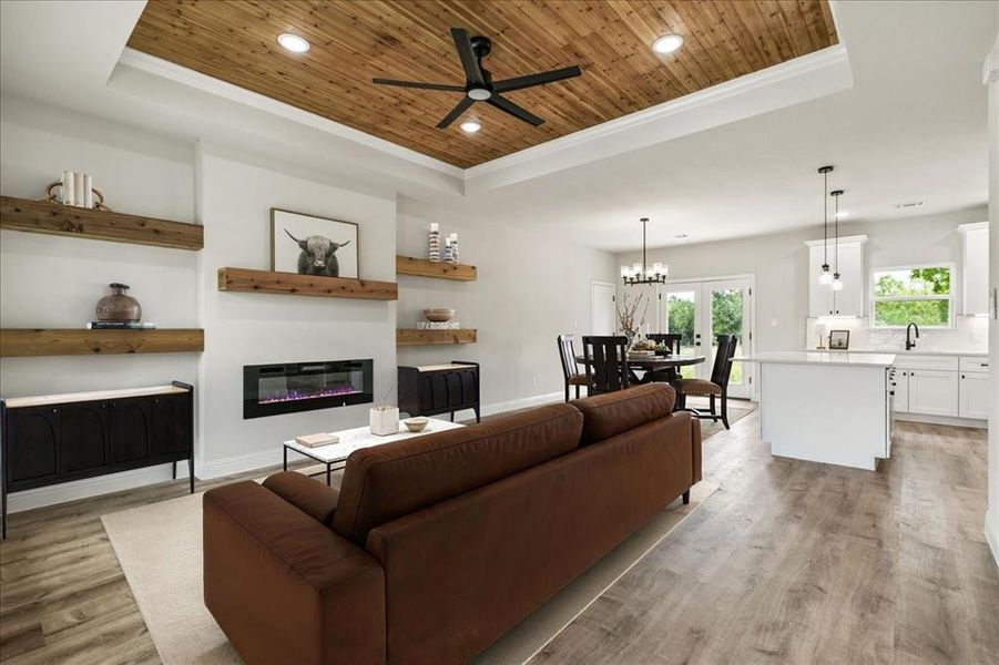 Open-concept living space featuring wood-finish flooring, a tray ceiling with wood paneling and recessed lighting, and an electric fireplace with floating wood mantle