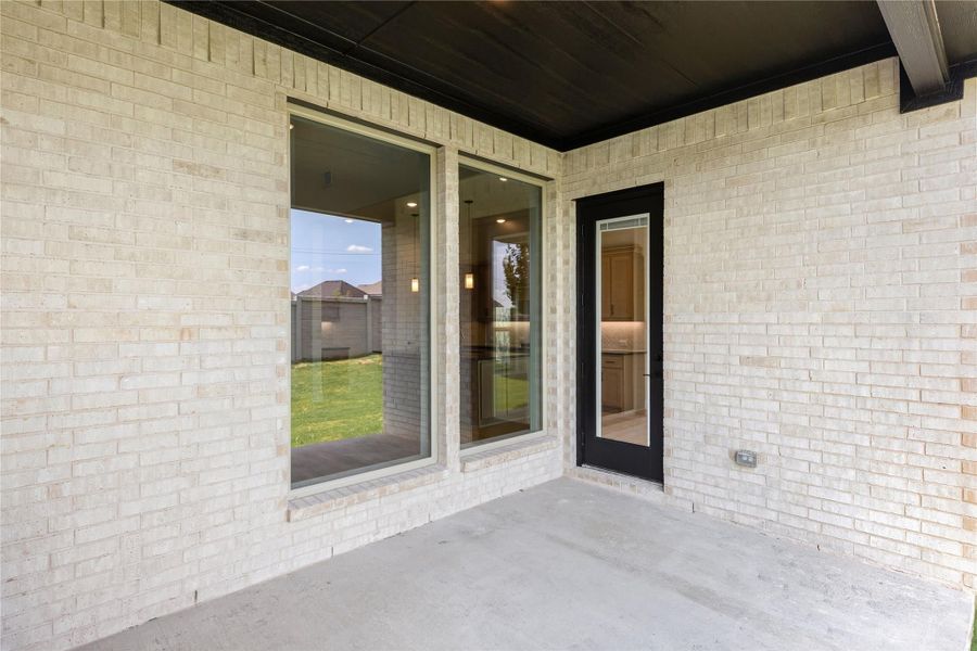 Covered concrete patio with light brick exterior, large windows, and black-framed back door leading inside.