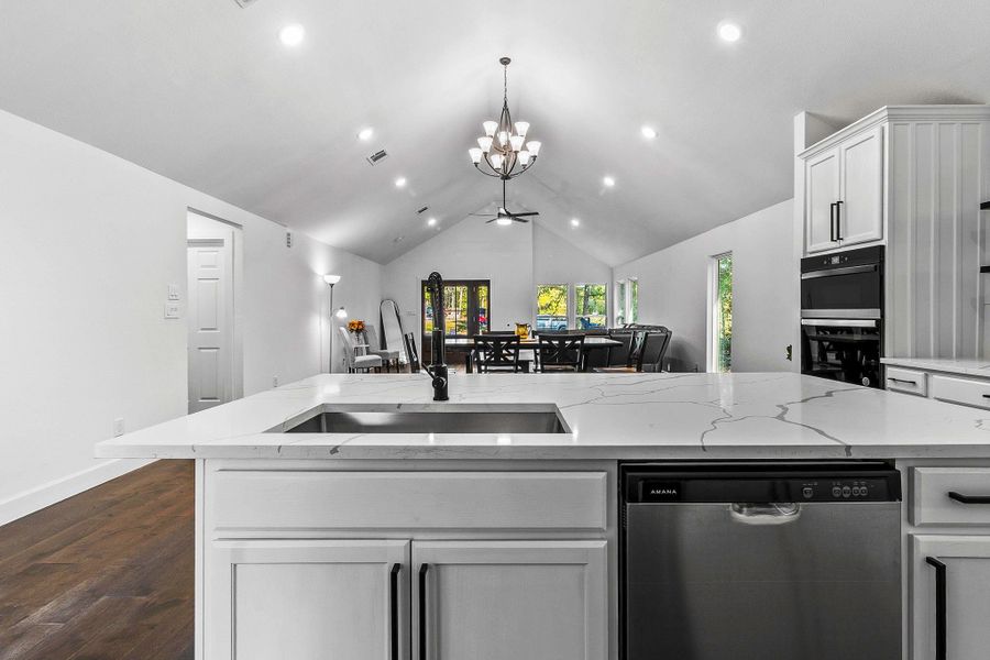 This photo showcases a modern, open-concept kitchen and living area with high ceilings and recessed lighting. The kitchen features sleek white cabinetry, a large island with a quartz countertop, and stainless steel appliances. The space is bright and airy, leading into a dining area with large windows offering natural light and views of greenery.
