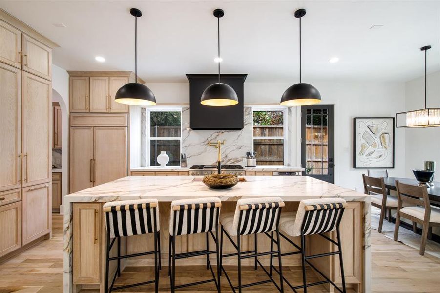 Open-concept kitchen featuring an oversized island with a waterfall countertop, integrated paneled refrigerator, built-in cabinetry, wood-finish flooring, and pendant lighting Open-concept kitchen featuring an oversized island with a waterfall countertop, integrated paneled refrigerator, built-in cabinetry, wood-finish flooring, and pendant lighting