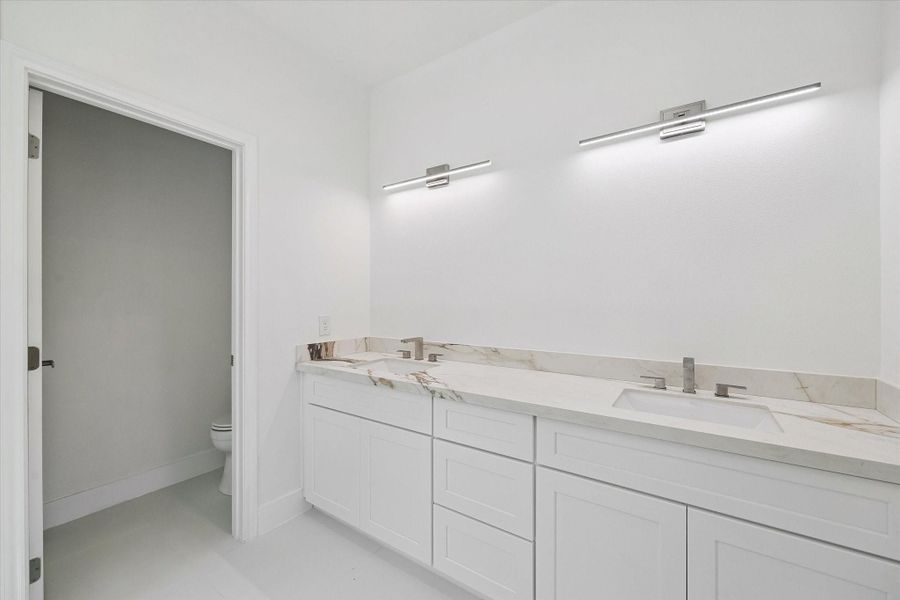 A spacious double vanity adds everyday convenience to the primary bath, featuring two under-mount sinks, sleek modern faucets, abundant cabinetry, and an expansive marble-look countertop. Contemporary linear lighting creates a streamlined appearance, while the bright white finishes keep the space feeling fresh and open. The adjacent private water closet adds another layer of functionality.