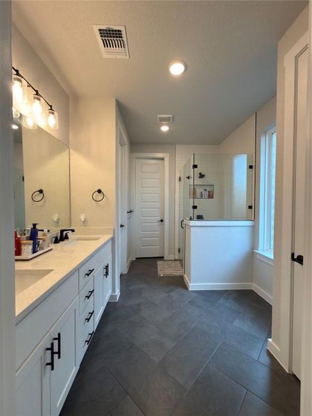 Spacious bathroom featuring a dual vanity with white cabinetry and light countertops, a glass-enclosed shower, dark tile flooring in a herringbone pattern, and recessed lighting throughout