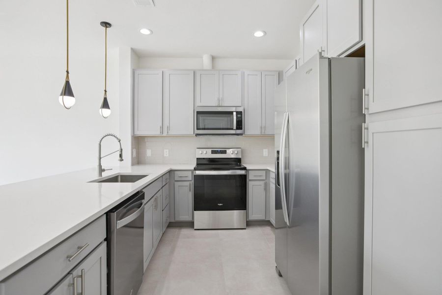 Kitchen with stainless steel appliances, hanging light fixtures, gray cabinets, light stone countertops, and backsplash