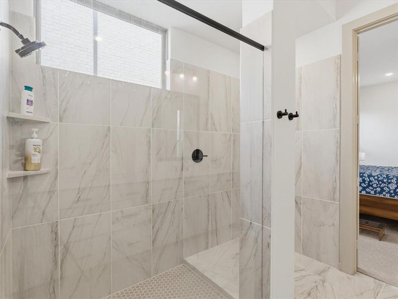 Walk-in shower featuring large format marbled tile, built-in shelving, a shower bench, hexagonal mosaic floor tile, and a black-finish shower head and controls