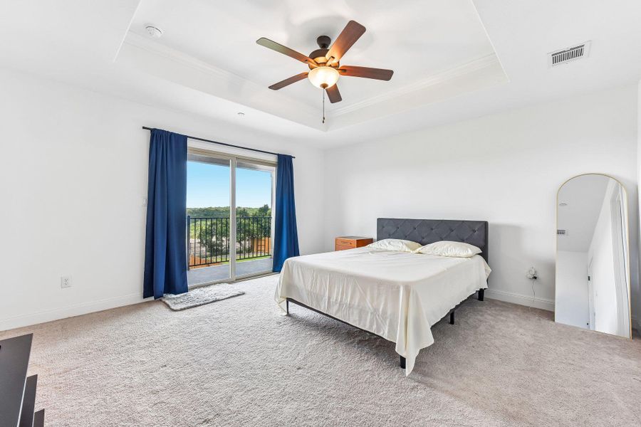 A spacious primary bedroom features a private balcony overlooking verdant scenery, with a tray ceiling and a ceiling fan.