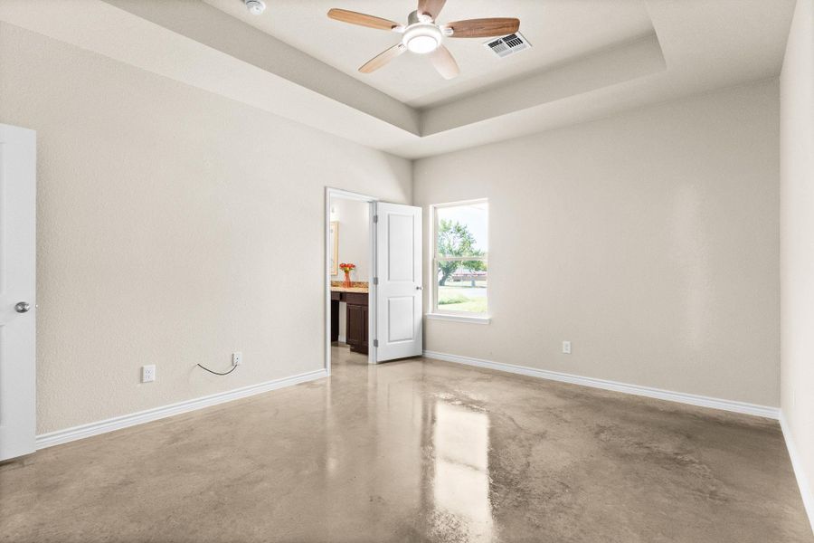 Unfurnished bedroom with finished concrete floors, a tray ceiling, connected bathroom, and a ceiling fan