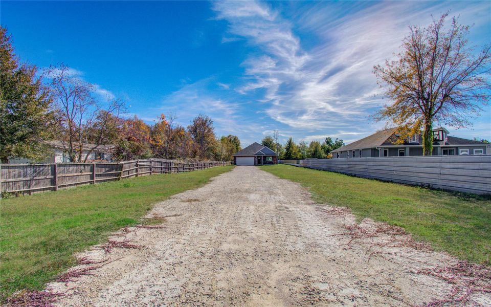 This photo shows a long gravel driveway leading to a detached garage or outbuilding. It's flanked by fenced grassy areas and neighboring homes, with a clear blue sky above. Ideal for buyers seeking space and privacy.