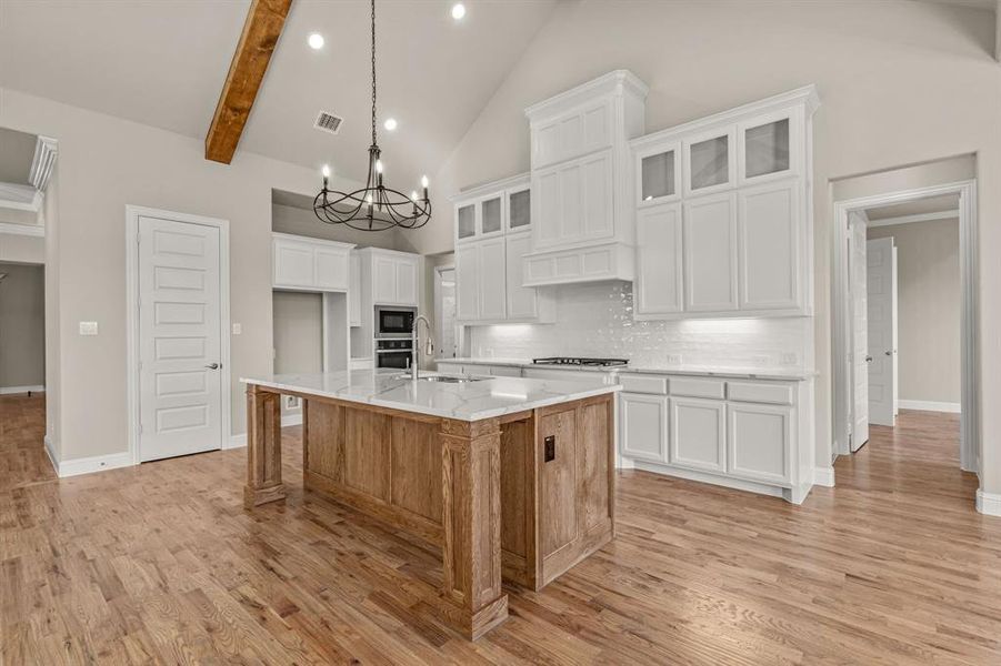 Kitchen featuring white cabinetry, backsplash, light stone countertops, a center island with sink, and beam ceiling