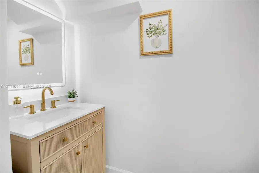 Fluted wood-finish vanity with a stone countertop, integrated rectangular sink, brass-toned faucet, and an LED-backlit mirror Fluted wood-finish vanity with a stone countertop, integrated rectangular sink, brass-toned faucet, and an LED-backlit mirror