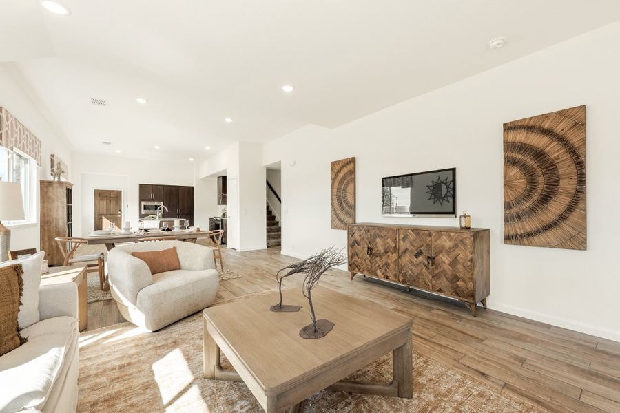 Open-concept living room with wood floors, white seating, and kitchen visible in background