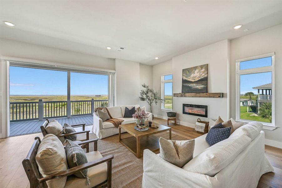 Walls of disappearing 16 foot sliding glass doors frame breathtaking bay views while filling the living room with natural light. White oak floors, soaring ceilings, and a sleek fireplace create a warm yet elevated space designed for entertaining and relaxed coastal living.