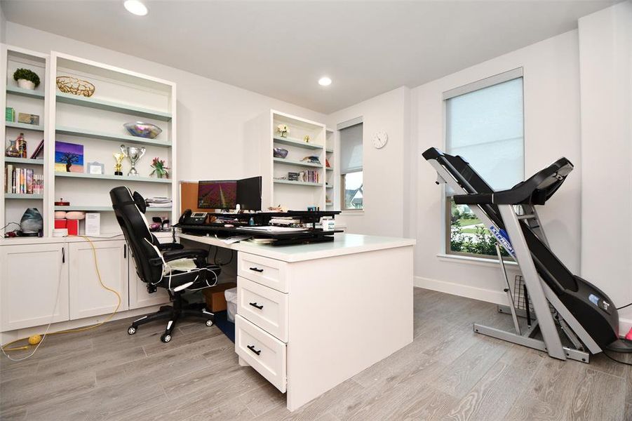 This bright home office features built-in shelving. The room has modern finishes, ample storage, and natural light from large windows, creating an inviting workspace.