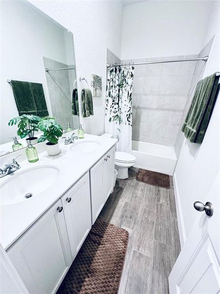 Full bathroom with toilet, wood-type flooring, shower / bath combination with curtain, and double vanity