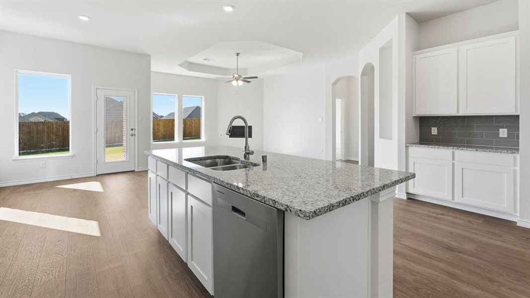 Open concept living space featuring a kitchen island with a double basin sink and a stone countertop, white cabinetry with a grey subway tile backsplash, and wood-style flooring throughout Open concept living space featuring a kitchen island with a double basin sink and a stone countertop, white cabinetry with a grey subway tile backsplash, and wood-style flooring throughout