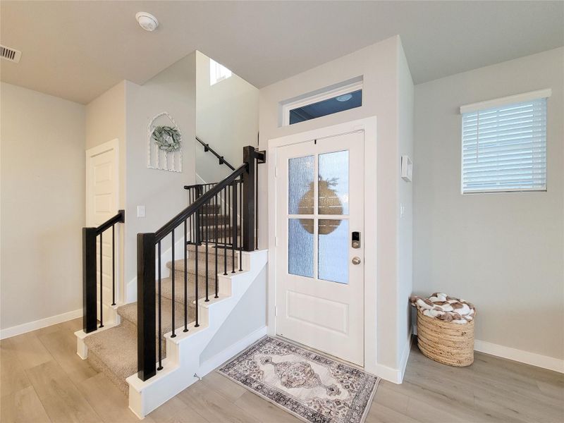 Welcoming entryway with a stylish front door featuring frosted glass panels. The area is bright, with light wood flooring and neutral walls. A carpeted staircase with black railings leads to the upper floor, adding a touch of elegance.