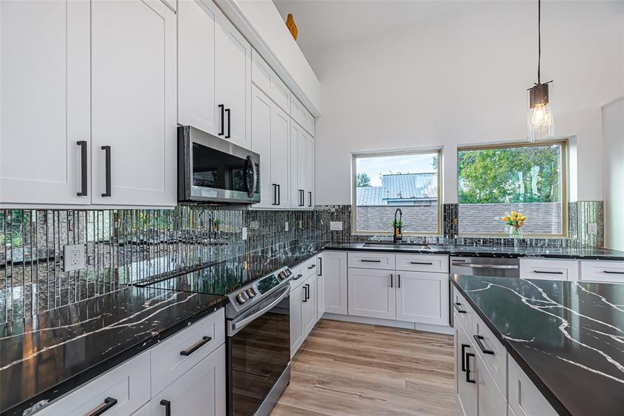 Modern kitchen featuring white cabinetry, sleek black quartz countertops with white veining, mosaic mirror tile backsplash, stainless steel appliances, and pendant lighting. Natural light flows through two windows overlooking greenery outside.