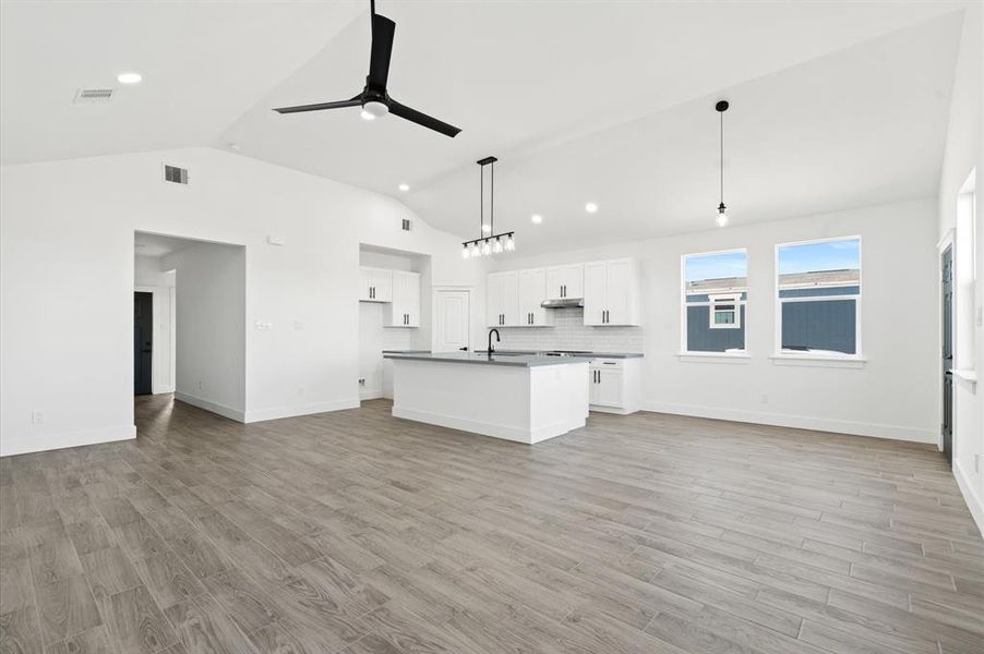 This is a spacious, modern open-plan living space with a kitchen, featuring white cabinetry, sleek countertops, stainless steel appliances, and a central island. The area is well-lit with natural light from large windows and complemented by contemporary light fixtures and a ceiling fan. The flooring is a light wood-look tile, providing a clean and airy feel.