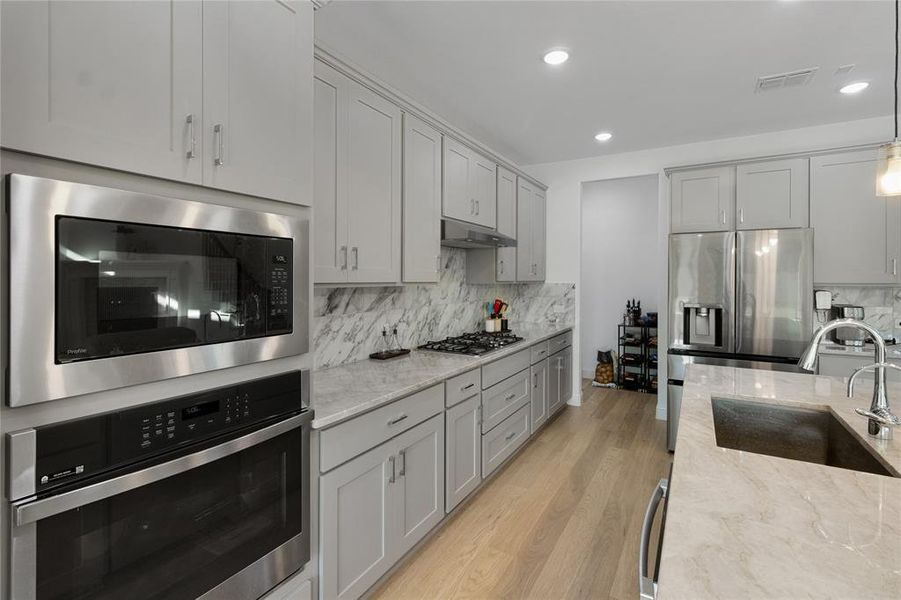 Modern kitchen featuring light gray cabinetry, stainless steel appliances, marble-look countertops, a built-in oven, and wood-finish flooring