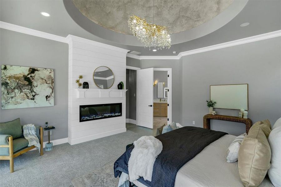 Carpeted bedroom with a fireplace, a notable chandelier, a tray ceiling, connected bathroom, and ornamental molding