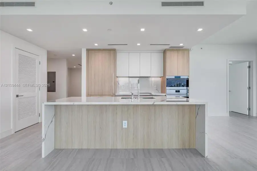 Kitchen featuring a center island with a stone-finish waterfall countertop, custom wood-finish cabinetry, white matte upper cabinets, integrated stainless steel appliances, and neutral flooring