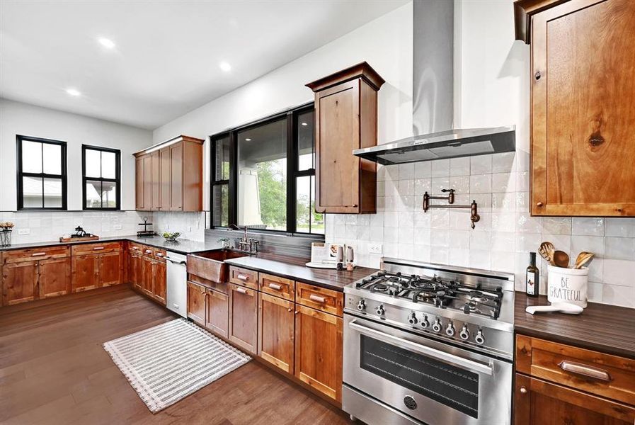 Kitchen with stainless steel appliances, wall chimney range hood, tasteful backsplash, dark wood-style floors, and brown cabinetry