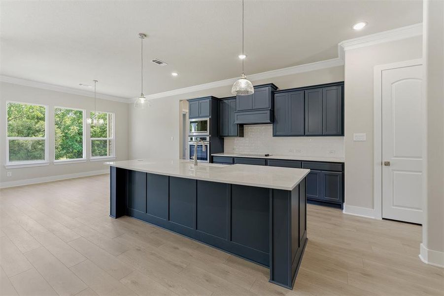 Open-concept kitchen featuring a spacious island with a light-toned countertop, dark-toned cabinetry, stainless steel built-in appliances, and light wood-finish flooring
