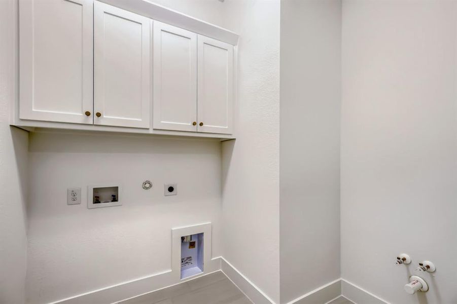 Laundry area featuring shaker-style upper cabinetry with brass-toned hardware, white walls, and light-toned tile flooring Laundry area featuring shaker-style upper cabinetry with brass-toned hardware, white walls, and light-toned tile flooring
