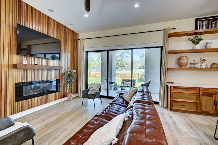 This cozy living room features a stylish wood-paneled wall with a modern fireplace and mounted TV. It offers natural light through large sliding glass doors leading to a patio. The space includes built-in shelves anproviding comfort and functionality.