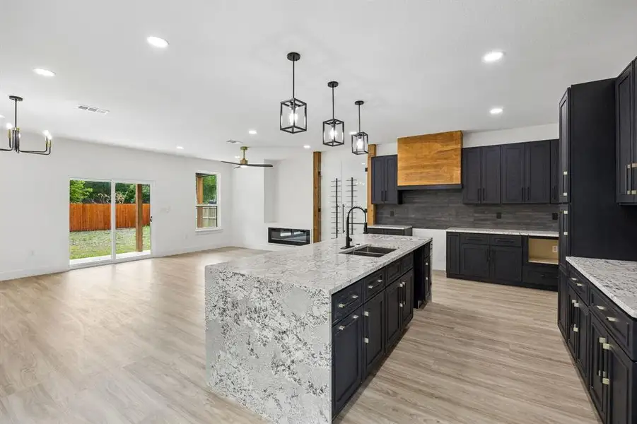 Open-concept living space featuring a spacious kitchen island with a waterfall countertop, dark cabinetry, and a wood-finish range hood Open-concept living space featuring a spacious kitchen island with a waterfall countertop, dark cabinetry, and a wood-finish range hood