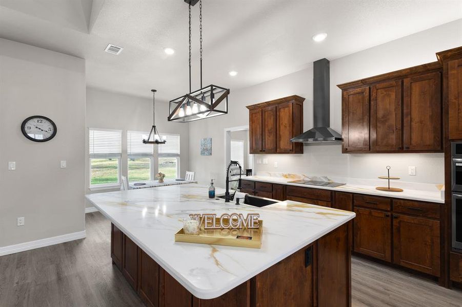 Kitchen featuring an island with sink, light stone counters, wood finished floors, hanging light fixtures, and wall chimney exhaust hood
