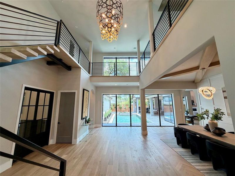 Two-story foyer featuring light wood-finish flooring, a contemporary chandelier, open-tread staircase with metal railing, and floor-to-ceiling glass doors opening to a backyard pool Two-story foyer featuring light wood-finish flooring, a contemporary chandelier, open-tread staircase with metal railing, and floor-to-ceiling glass doors opening to a backyard pool
