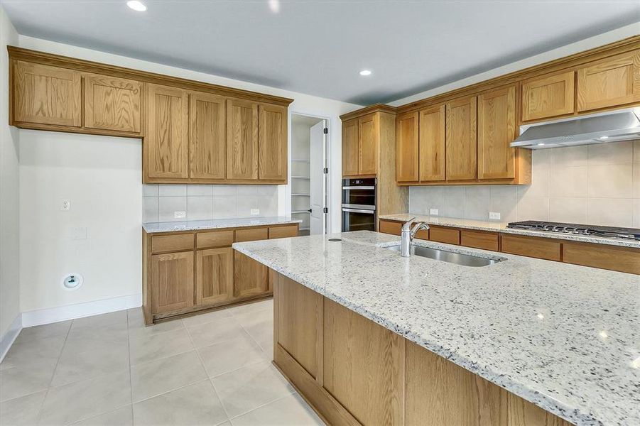 Kitchen featuring light wood cabinetry, light-toned countertops, a stainless steel sink with a gooseneck faucet, a built-in double oven, and a gas range with an overhead vent hood Kitchen featuring light wood cabinetry, light-toned countertops, a stainless steel sink with a gooseneck faucet, a built-in double oven, and a gas range with an overhead vent hood