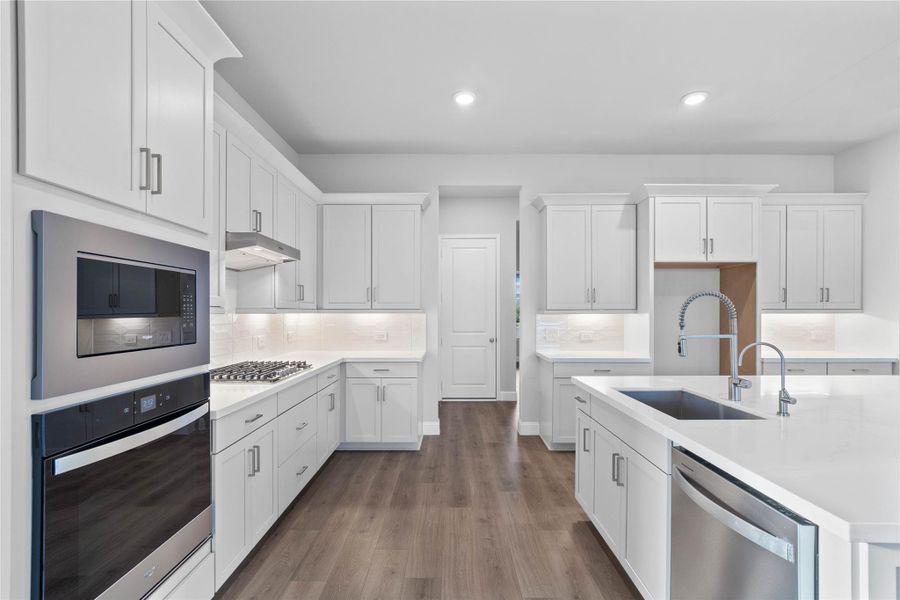 This gourmet kitchen is a study in modern elegance, featuring a cohesive design of crisp white Shaker-style cabinetry, sleek quartz countertops, and a seamless white subway tile backsplash. The chef-inspired space is fully equipped with high-end stainless steel appliances, including a built-in double oven and a gas cooktop, all anchored by a large central island with a deep undermount sink and industrial-style pull-down faucet.