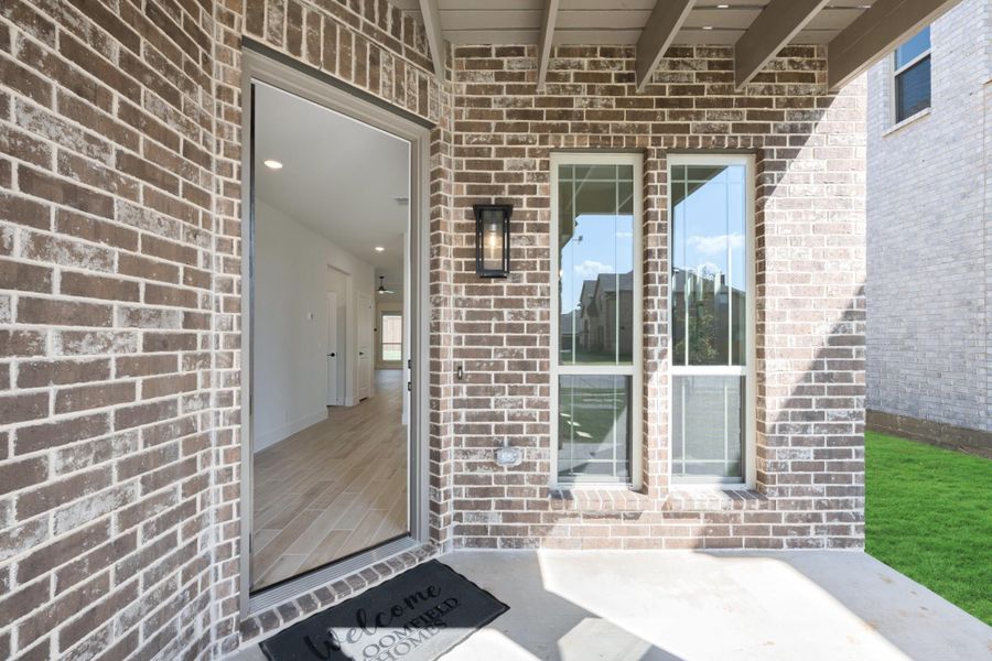 Home entrance with brick exterior, open front door, wall lantern, and welcome mat on covered porch